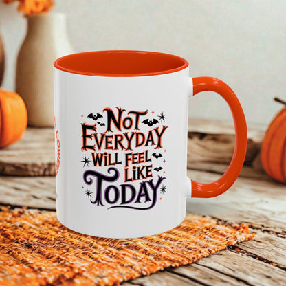 A white ceramic mug with a bright orange handle and interior, featuring the printed phrase 'Not Everyday Will Feel Like Today' in decorative black and orange lettering with small bat and star accents. The mug sits on a woven orange placemat on a rustic wooden surface, with pumpkins and blurred decor in the background.