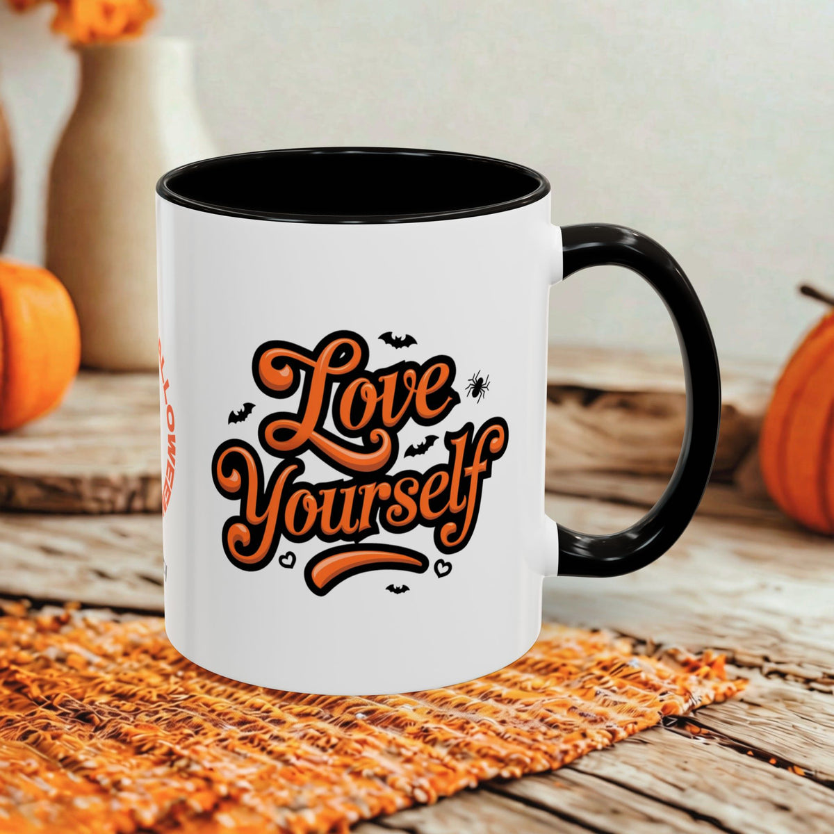 A white ceramic coffee mug with a black handle and interior, featuring the phrase 'Love Yourself' in orange script with small black bat and spider graphics. The mug is styled on a woven placemat over a rustic wooden table with pumpkins in the background.