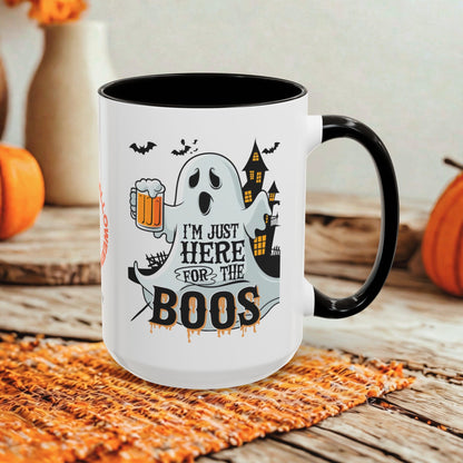 A white ceramic mug with a black handle and interior, printed with a Halloween graphic featuring a ghost holding a beer mug, bats, a haunted house, and the text 'I'M JUST HERE FOR THE BOOS'. The mug is set on a woven orange placemat on a rustic wooden table, with pumpkins and blurred decorative vases in the background.