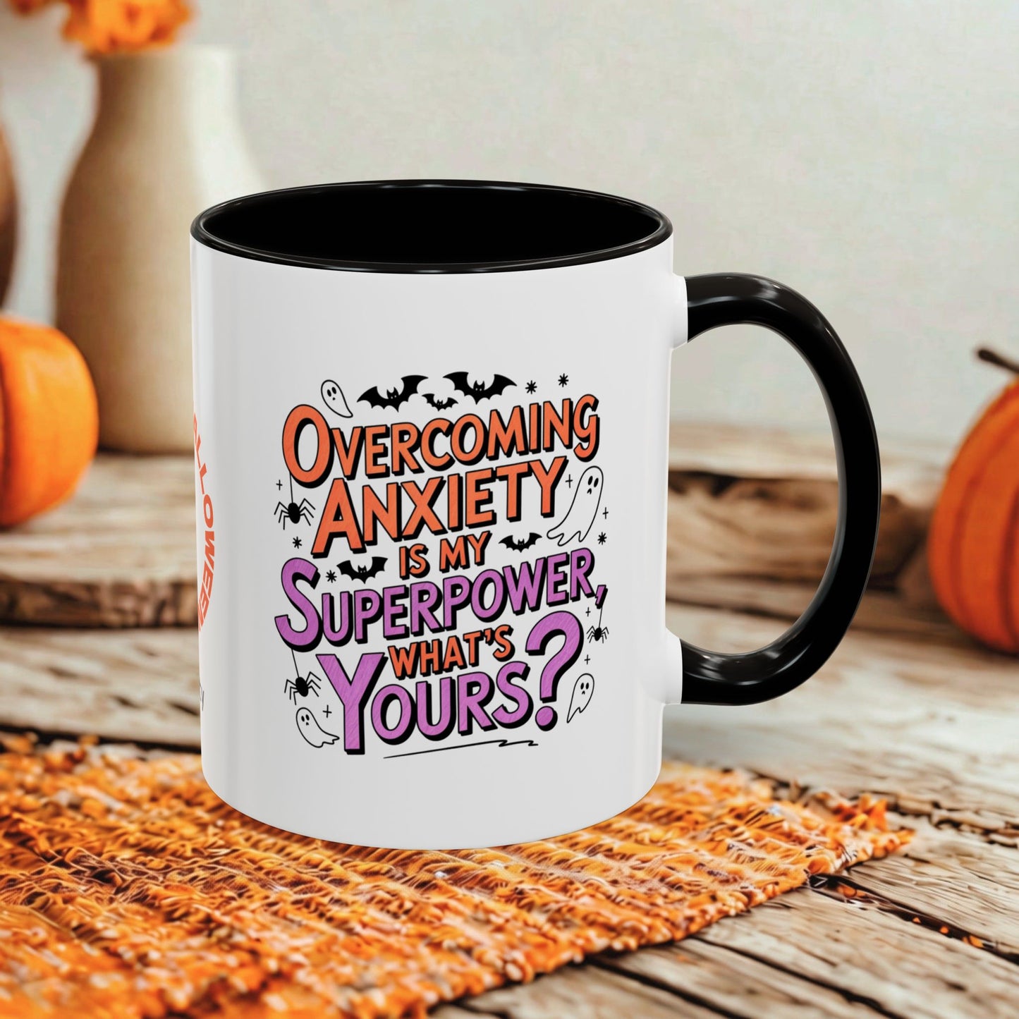 A white ceramic mug with a black handle and interior, printed with the phrase 'Overcoming Anxiety is my Superpower, What's Yours?' in orange and purple lettering. The mug is styled on a rustic wooden surface with a woven orange placemat and blurred pumpkins in the background.