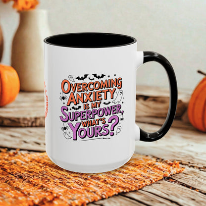 A white ceramic mug with a black handle and interior, featuring the printed text 'Overcoming Anxiety is my Superpower, what's yours?' in orange and purple lettering. The mug is styled on a rustic wooden surface with a woven orange placemat and blurred orange pumpkins in the background.