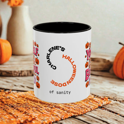 A white ceramic coffee mug with a black interior, featuring a personalized Halloween design with orange and black text and pumpkin illustrations. The mug is set on a rustic wooden table with autumn-themed props like pumpkins and a woven placemat.