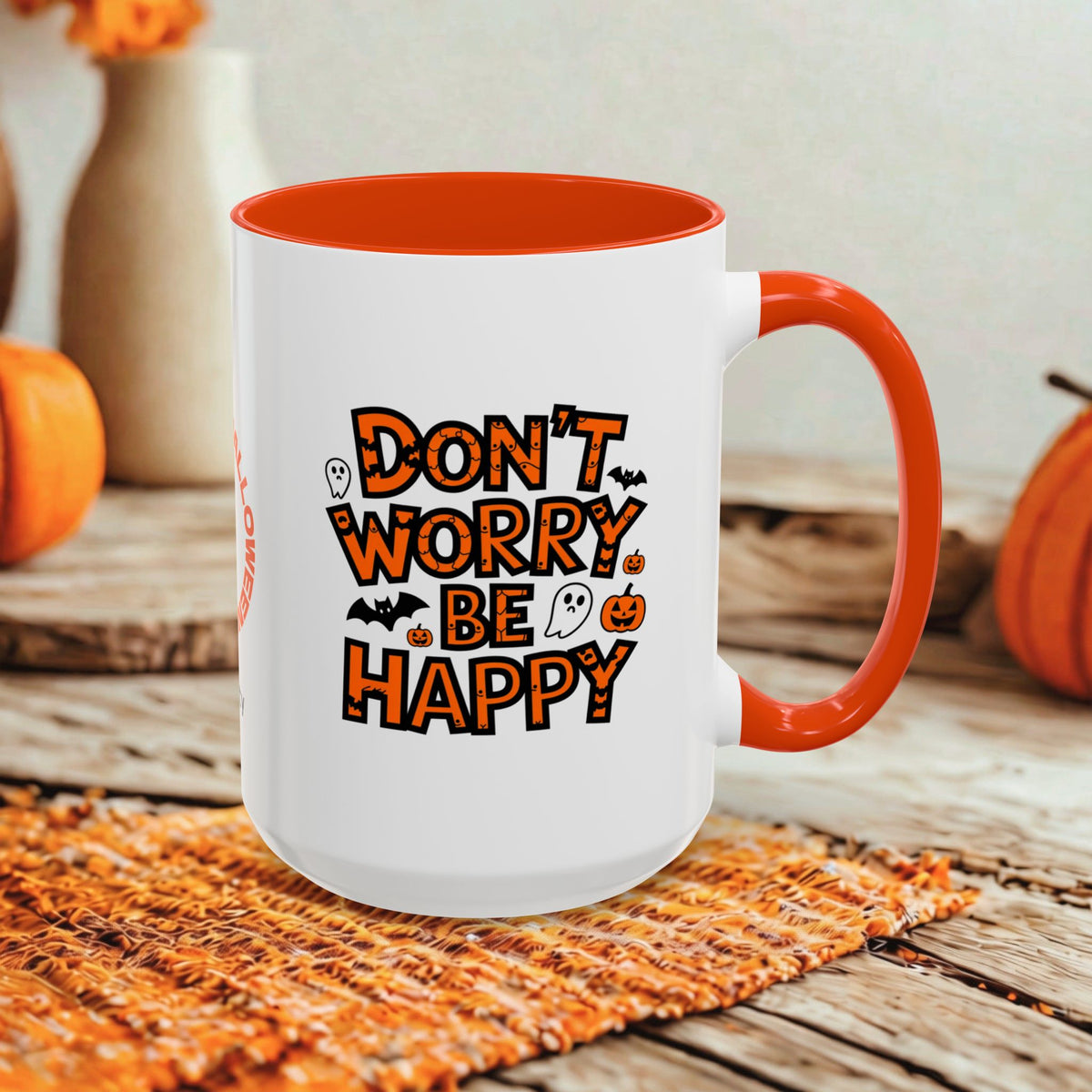 A white ceramic mug with a bright orange handle and interior, featuring the phrase 'DON'T WORRY BE HAPPY' in orange lettering with small Halloween-themed graphics. The mug is styled on a rustic wooden surface with autumn props like pumpkins and a woven placemat.