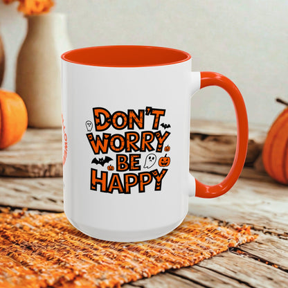 A white ceramic mug with a bright orange handle and interior, featuring the phrase 'DON'T WORRY BE HAPPY' in orange lettering with small Halloween-themed graphics. The mug is styled on a rustic wooden surface with autumn props like pumpkins and a woven placemat.