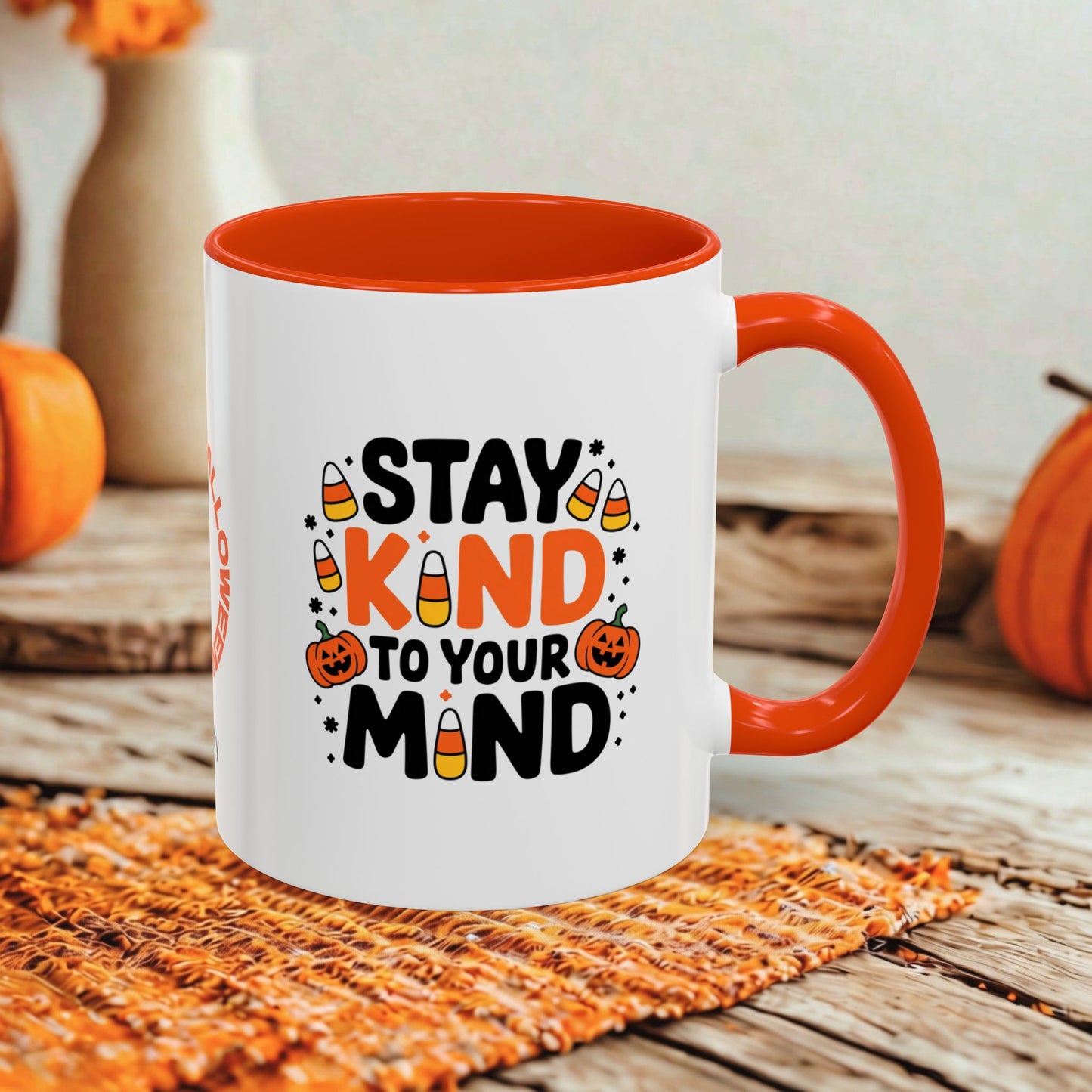 A white ceramic mug with a bright orange interior, handle, and rim, featuring a Halloween-themed graphic that reads 'STAY KIND TO YOUR MIND' in black and orange lettering with candy corn and jack-o'-lantern illustrations. The mug is set on a woven orange placemat on a rustic wooden table, with blurred pumpkins and a vase in the background.