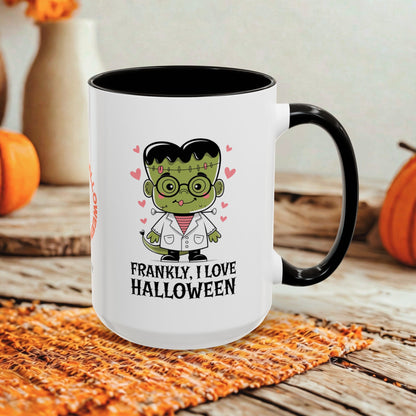 A white ceramic mug with a black handle and interior, featuring a cartoon Frankenstein character and the text 'FRANKLY, I LOVE HALLOWEEN' on the front. The mug is styled on a rustic wooden surface with autumn props including pumpkins and a woven placemat.