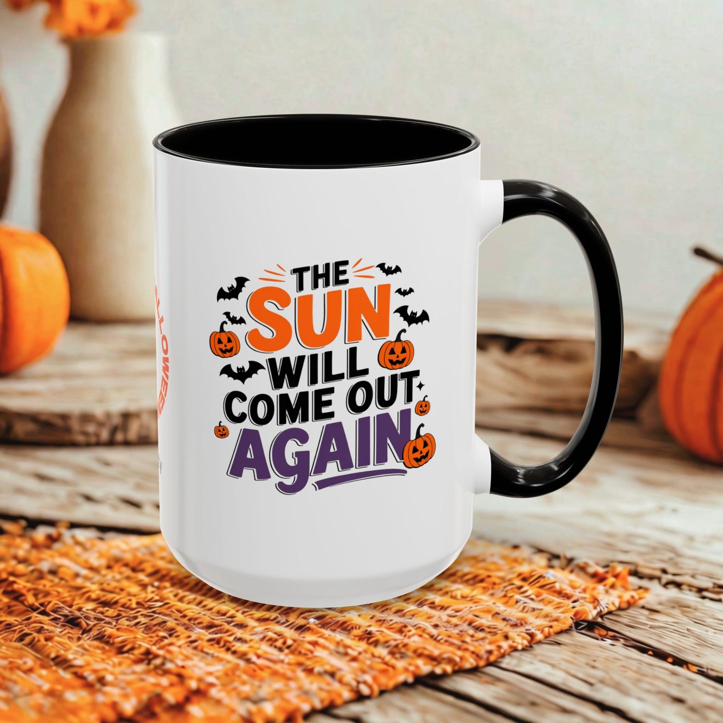 A white ceramic mug with a black handle and black interior, printed with the phrase 'THE SUN WILL COME OUT AGAIN' in orange, black, and purple lettering with Halloween-themed graphics. The mug is styled on a woven placemat with pumpkins and a neutral background.