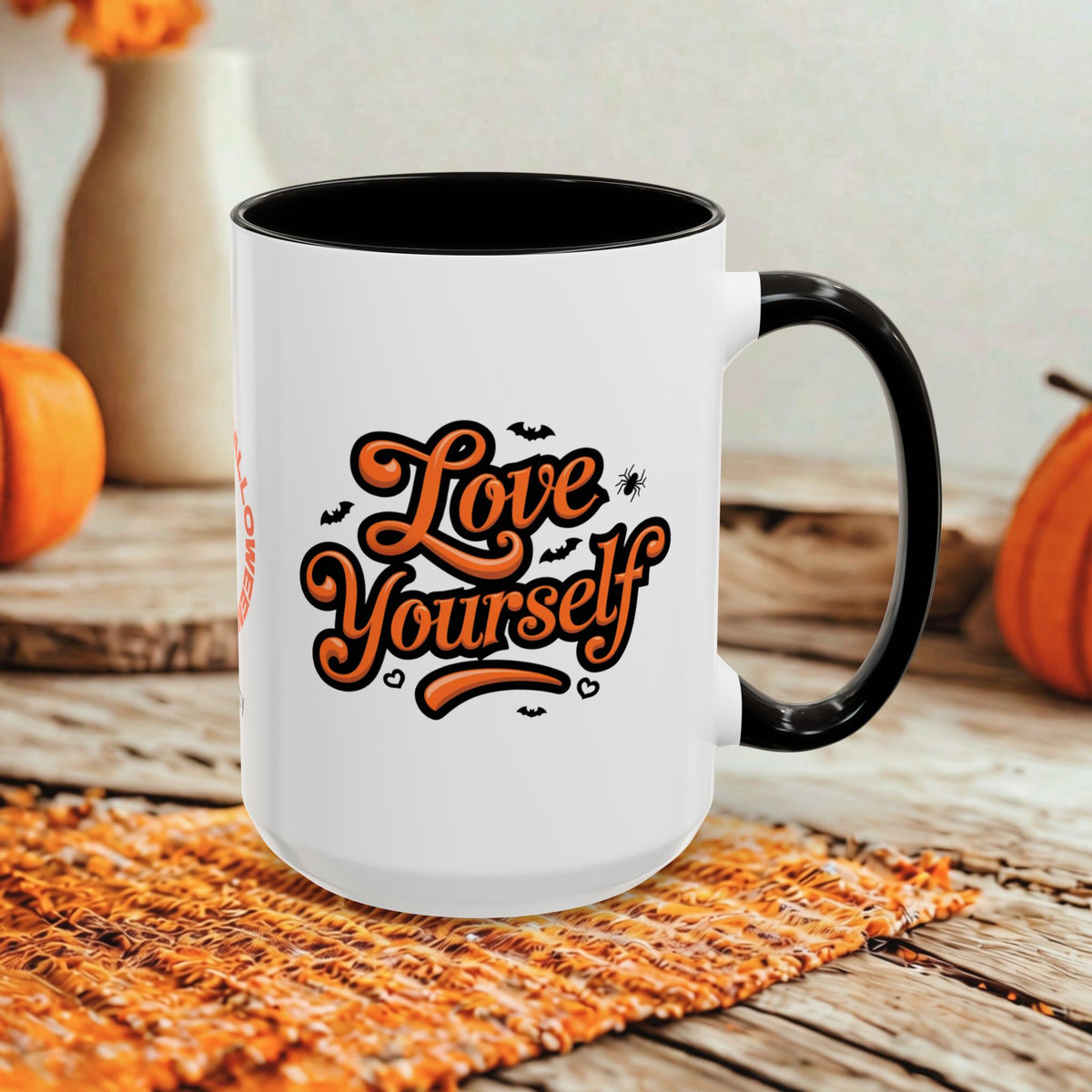 A white ceramic mug with a black handle and black interior, featuring the phrase 'Love Yourself' in orange script with small bat and spider graphics. The mug is styled on a woven orange placemat over a rustic wooden surface, with pumpkins and blurred decorative vases in the background.