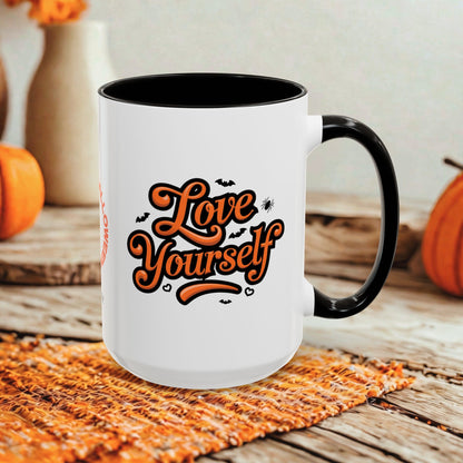 A white ceramic mug with a black handle and black interior, featuring the phrase 'Love Yourself' in orange script with small bat and spider graphics. The mug is styled on a woven orange placemat over a rustic wooden surface, with pumpkins and blurred decorative vases in the background.