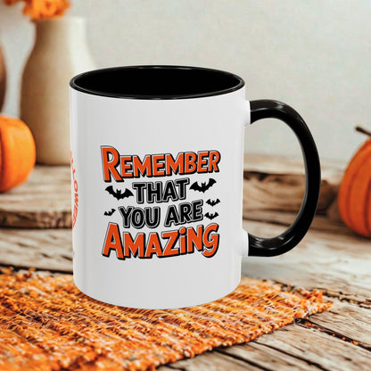 A white ceramic coffee mug with a black interior, handle, and rim, featuring the text 'REMEMBER THAT YOU ARE AMAZING' in orange and black with bat silhouettes. The mug is set on a woven orange placemat on a rustic wooden table, with pumpkins and a neutral background.