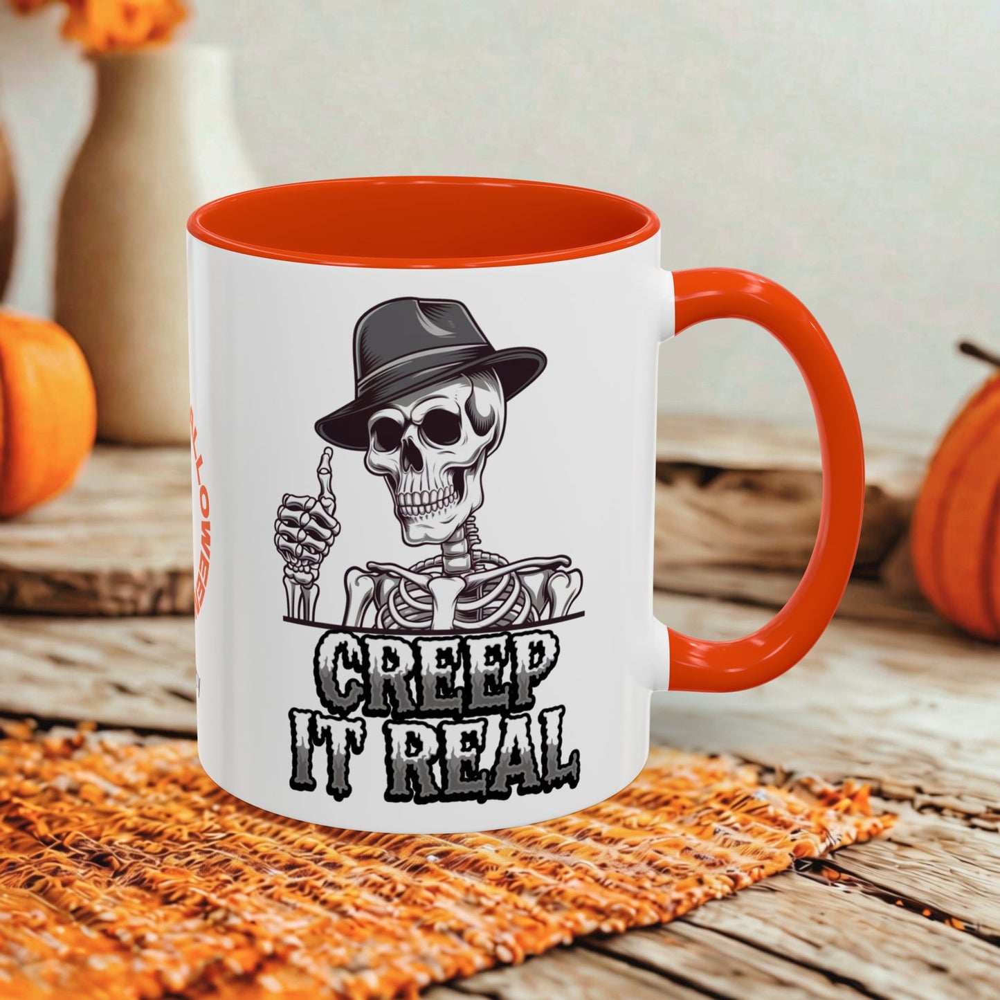 A white ceramic mug with a bright orange handle and interior, featuring a black-and-white skeleton graphic wearing a hat and the text 'CREEP IT REAL.' The mug is styled on a woven orange mat over a rustic wooden surface with pumpkins and a blurred background.