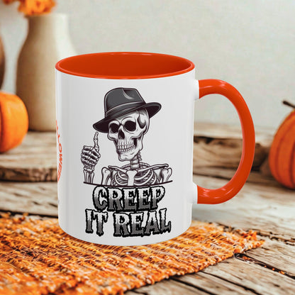 A white ceramic mug with a bright orange handle and interior, featuring a black-and-white skeleton graphic wearing a hat and the text 'CREEP IT REAL.' The mug is styled on a woven orange mat over a rustic wooden surface with pumpkins and a blurred background.