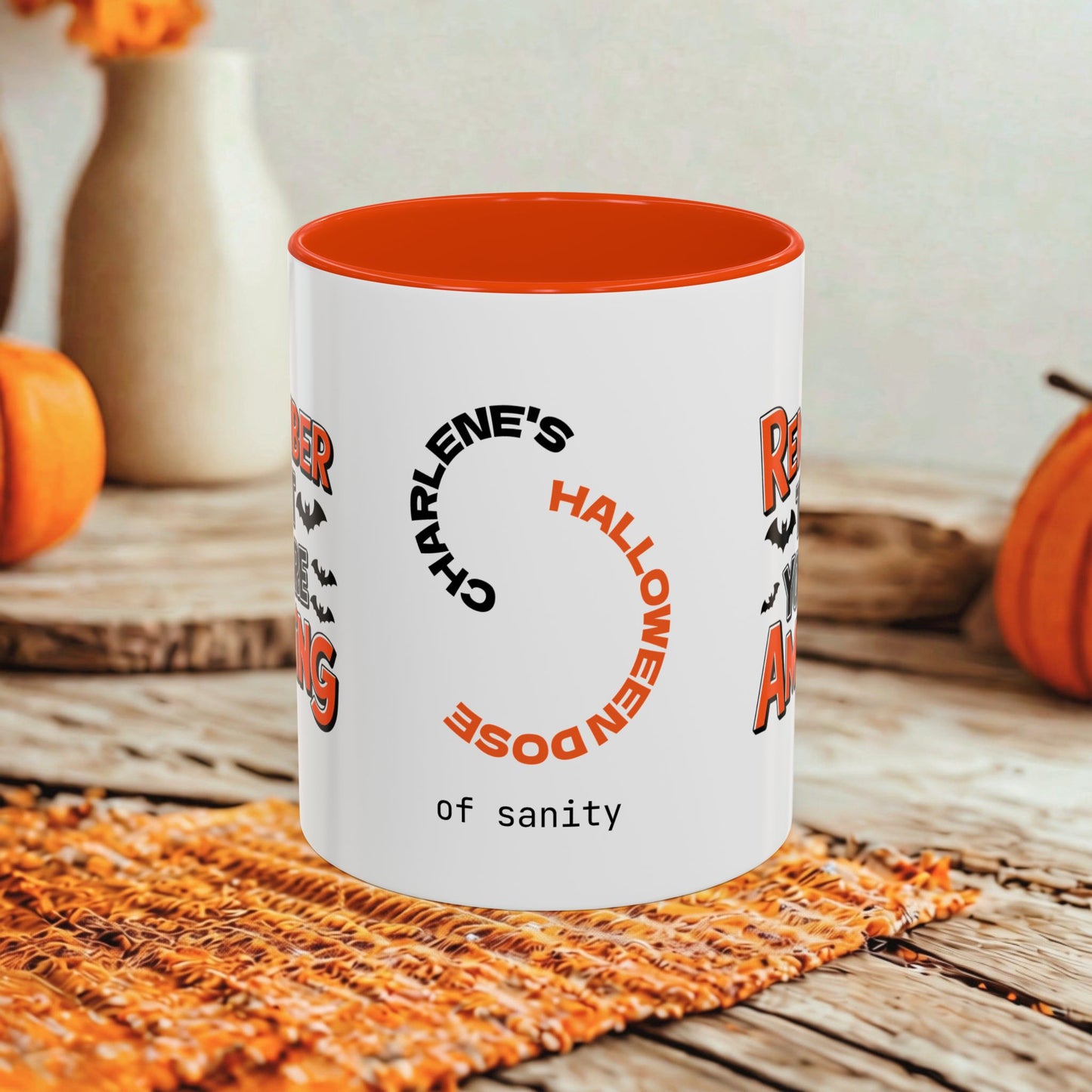 A white ceramic mug with a bright orange interior, featuring a Halloween-themed printed design with the text 'Charlene's Halloween Dose of sanity' in black and orange lettering. The mug is styled on a rustic wooden table with autumn props including pumpkins and a woven placemat.