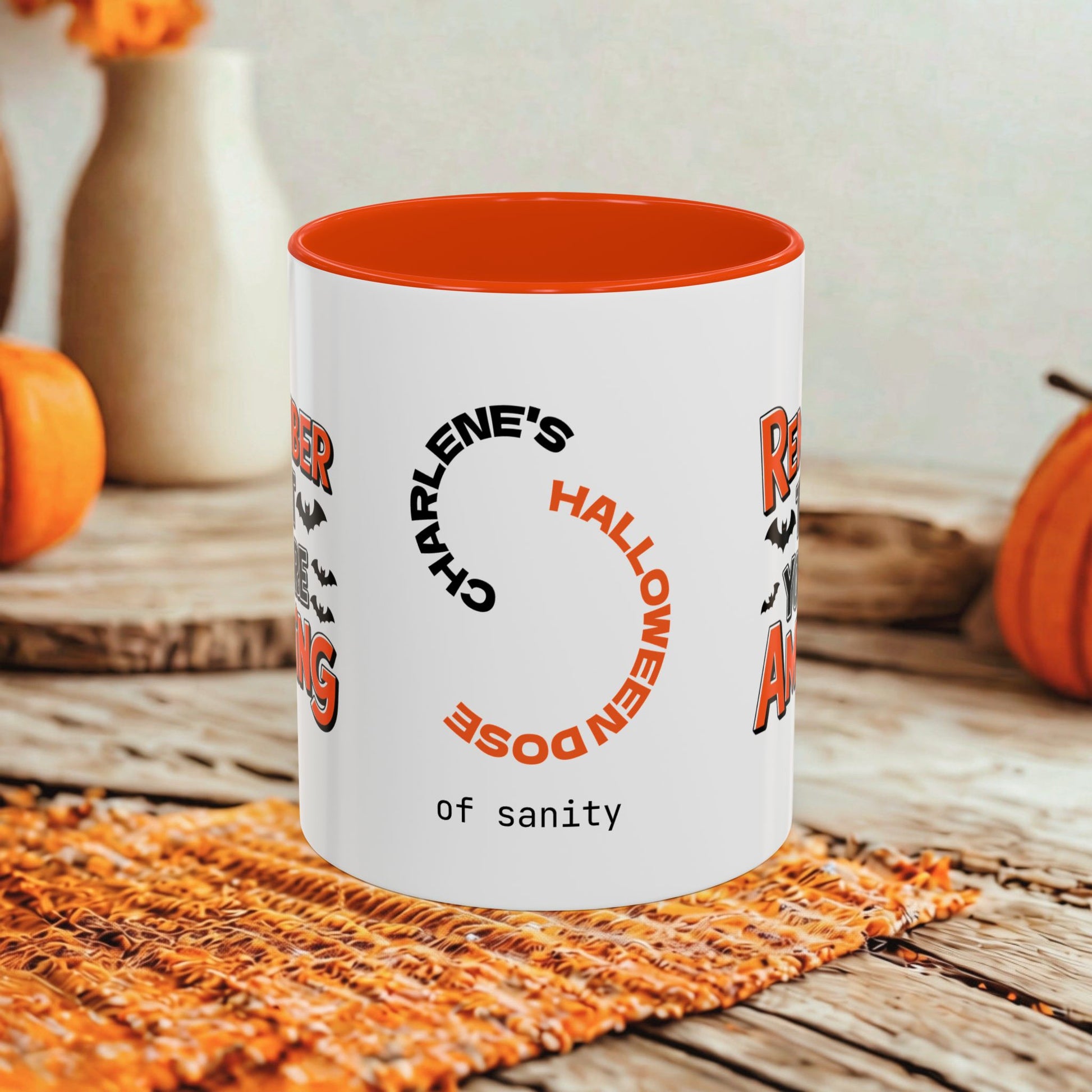 A white ceramic mug with a bright orange interior, featuring a Halloween-themed printed design with the text 'Charlene's Halloween Dose of sanity' in black and orange lettering. The mug is styled on a rustic wooden table with autumn props including pumpkins and a woven placemat.