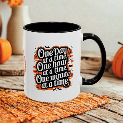 A white ceramic coffee mug with a black handle and interior, featuring a Halloween-themed graphic that reads 'One Day at a time, One hour at a time, One minute at a time.' The mug is styled on a rustic wooden table with a woven placemat and orange pumpkins in the background.