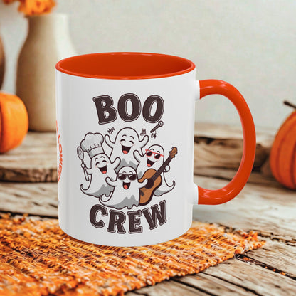 A white ceramic coffee mug with a bright orange handle and interior, featuring a cartoon graphic of four playful ghosts and the words 'BOO CREW' in dark brown lettering. The mug is styled on an orange woven placemat over a rustic wooden table, with decorative pumpkins and a blurred background.