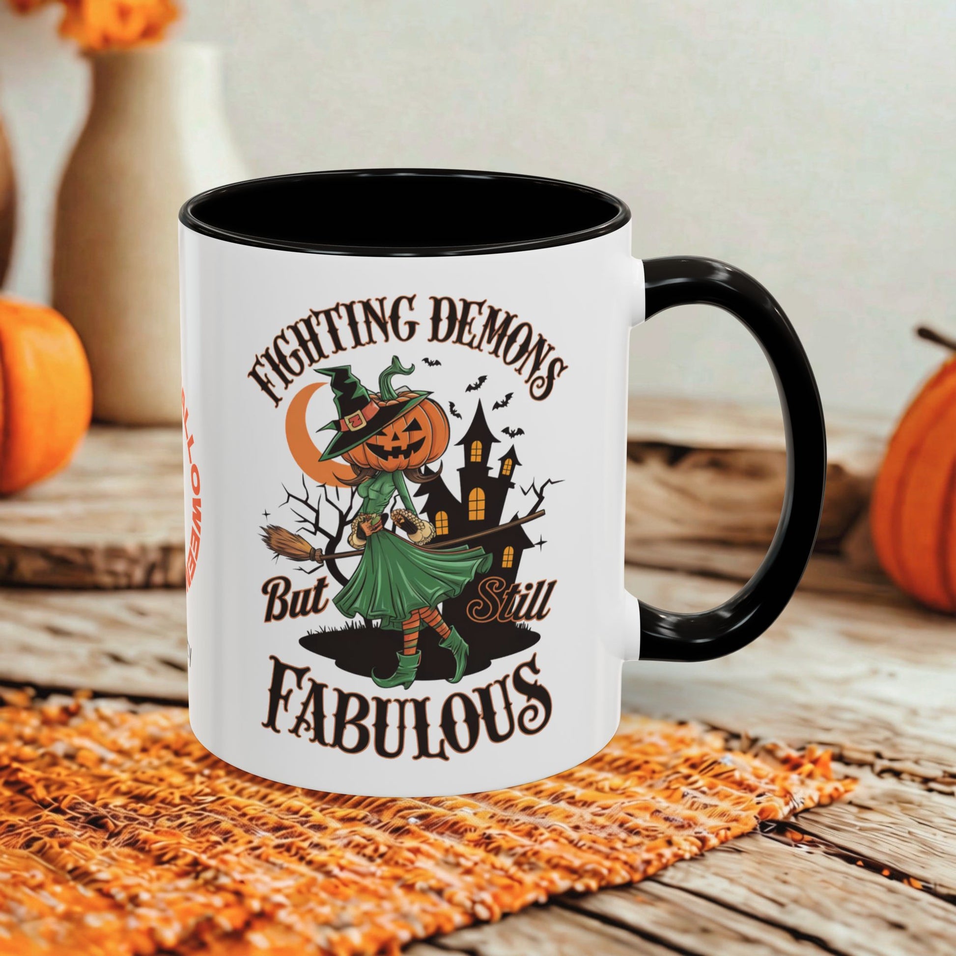 A white ceramic mug with a black handle and interior, featuring a Halloween-themed graphic of a witch with a pumpkin head, a haunted house, and the text 'Fighting Demons But Still Fabulous'. The mug is styled on a rustic wooden table with a woven orange placemat and autumn decorations in the background.