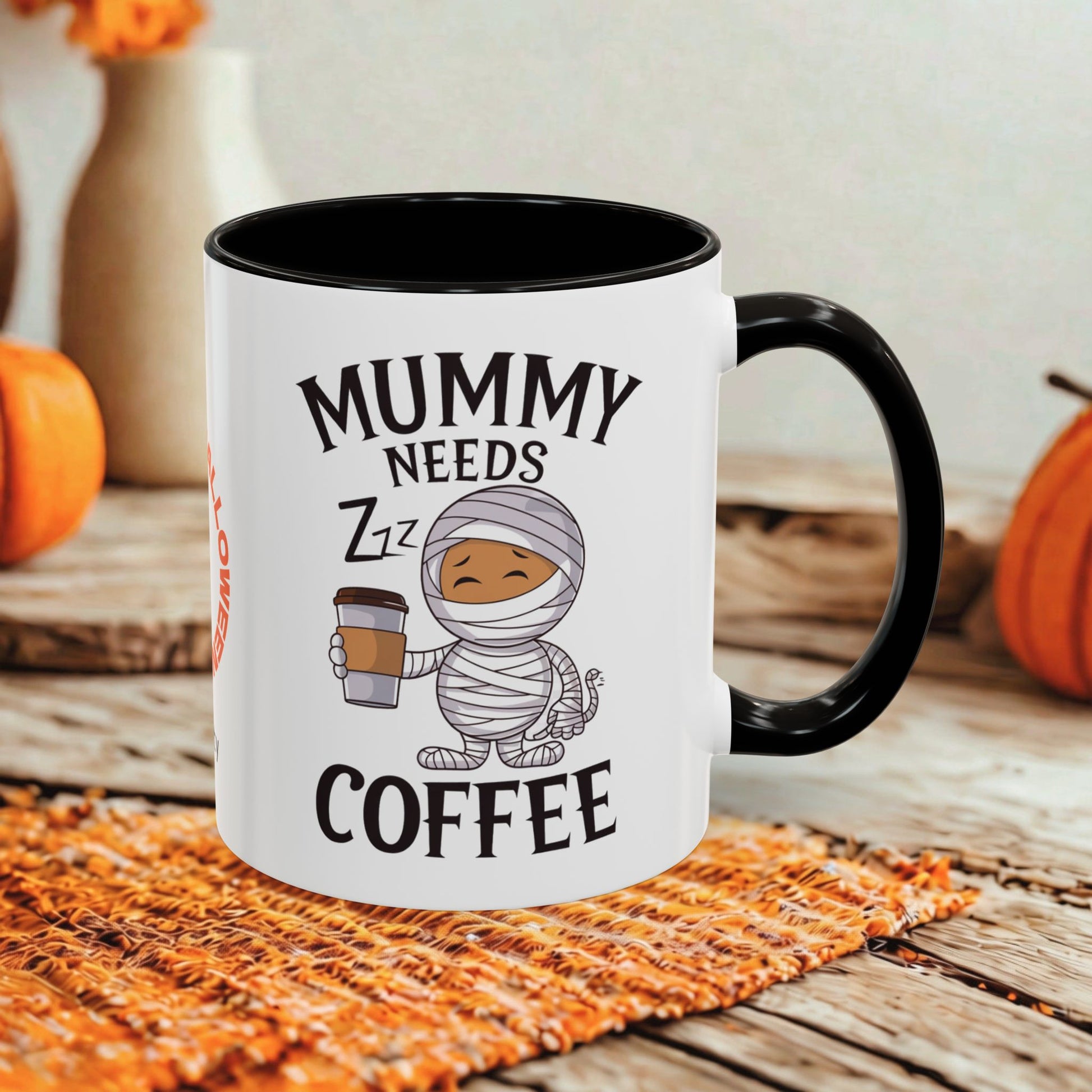 A white ceramic coffee mug with a black handle and interior, printed with the text 'MUMMY NEEDS COFFEE' and a cartoon mummy holding a takeaway coffee cup. The mug is shown on a woven orange placemat on a rustic wooden surface with orange pumpkins and a blurred background.