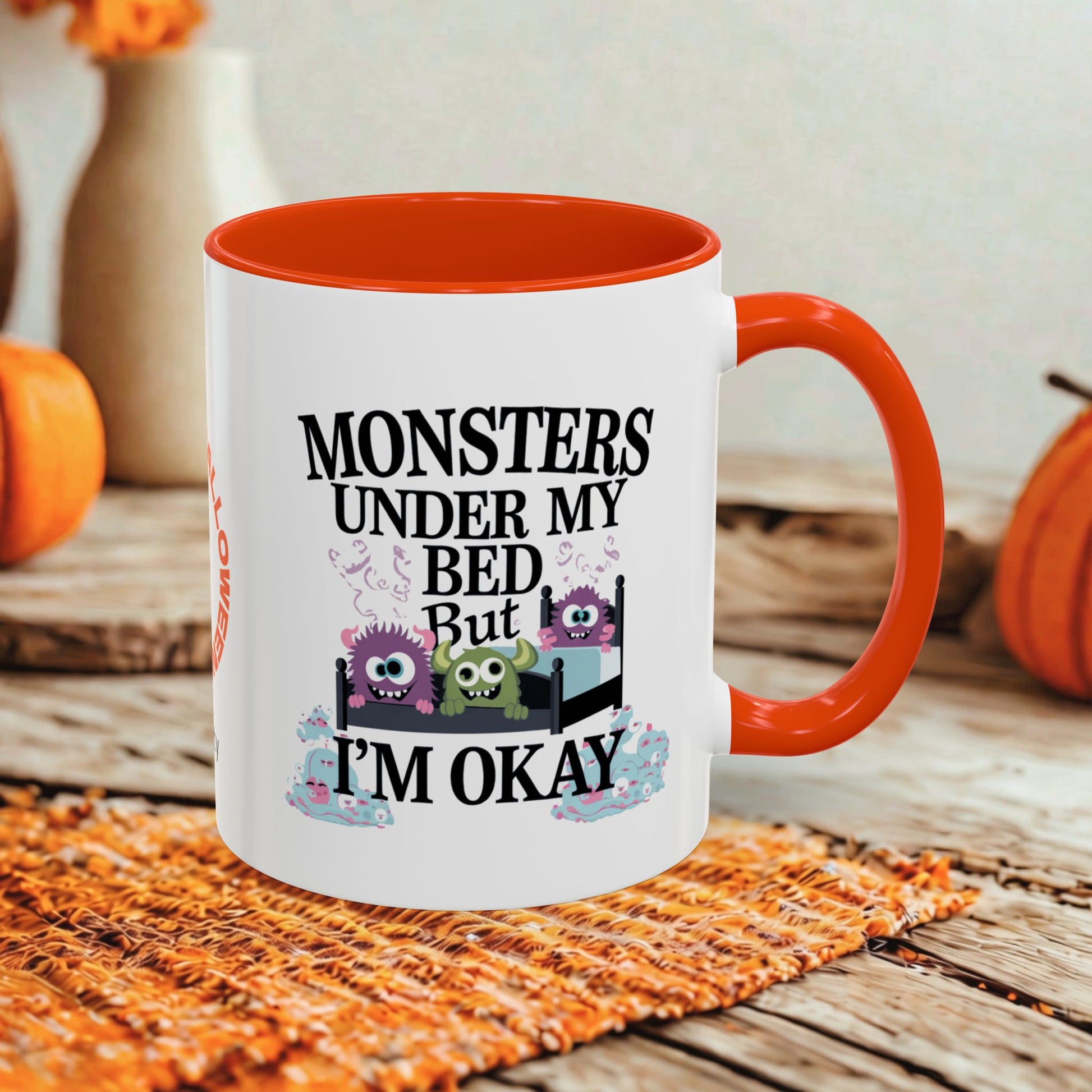 A white ceramic mug with a bright orange interior and handle, featuring a cartoon monster graphic and the text 'MONSTERS UNDER MY BED But I'M OKAY'. The mug is styled on an orange woven placemat over a rustic wooden table with pumpkins and a blurred background.