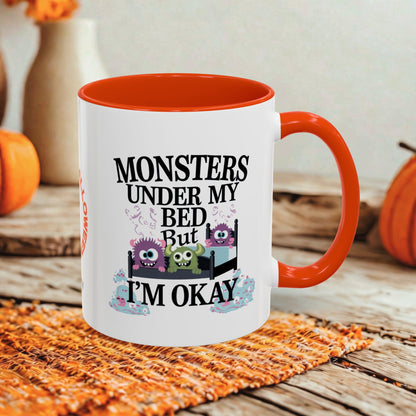 A white ceramic mug with a bright orange interior and handle, featuring a cartoon monster graphic and the text 'MONSTERS UNDER MY BED But I'M OKAY'. The mug is styled on an orange woven placemat over a rustic wooden table with pumpkins and a blurred background.