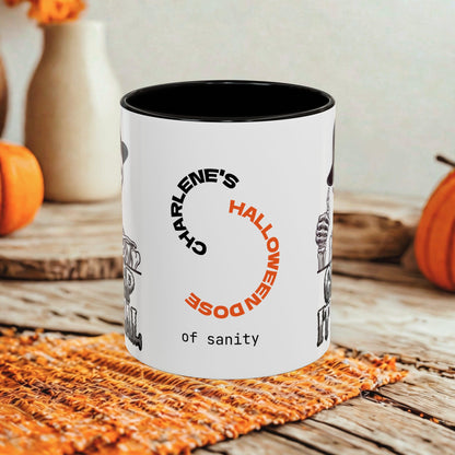 A white ceramic mug with a black interior and rim, printed with the phrase 'CHARLENE'S HALLOWEEN DOSE of SANITY' in black and orange text, styled on a rustic wooden surface with autumn props like pumpkins and a woven placemat.