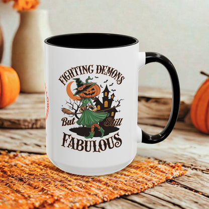 A white ceramic mug with a black handle and interior, featuring a Halloween-themed graphic that reads 'Fighting Demons But Still Fabulous' with an illustration of a witch, pumpkin, and haunted house. The mug is styled on a rustic wooden surface with autumn props in the background.