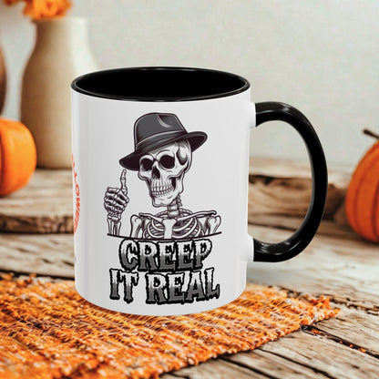 A white ceramic mug with a black handle and black interior, printed with a cartoon skeleton wearing a hat and the text 'CREEP IT REAL'. The mug is styled on a rustic wooden table with a woven placemat and seasonal pumpkins in the background.
