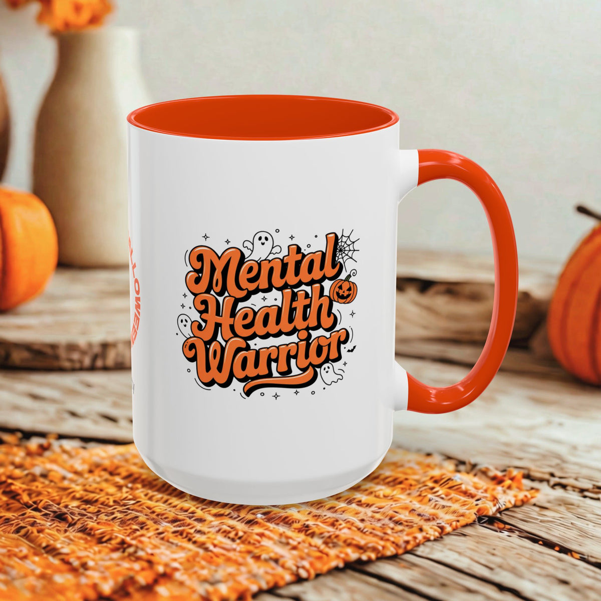 A white ceramic mug with a bright orange handle and interior, featuring the phrase 'Mental Health Warrior' in orange lettering with Halloween-themed doodles. The mug is styled on a woven orange placemat over a rustic wooden table, with pumpkins and a neutral background.
