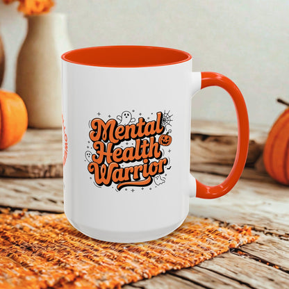 A white ceramic mug with a bright orange handle and interior, featuring the phrase 'Mental Health Warrior' in orange lettering with Halloween-themed doodles. The mug is styled on a woven orange placemat over a rustic wooden table, with pumpkins and a neutral background.