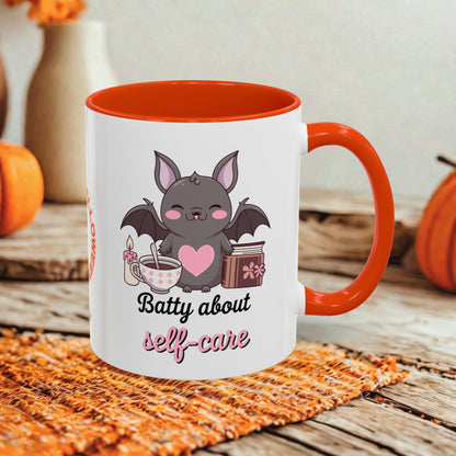 A white ceramic mug with a bright orange handle and interior, printed with a cute bat graphic and the phrase 'Batty about self-care' in black and pink script. The mug is styled on a woven orange placemat over a rustic wooden surface, with blurred pumpkins and a neutral background.