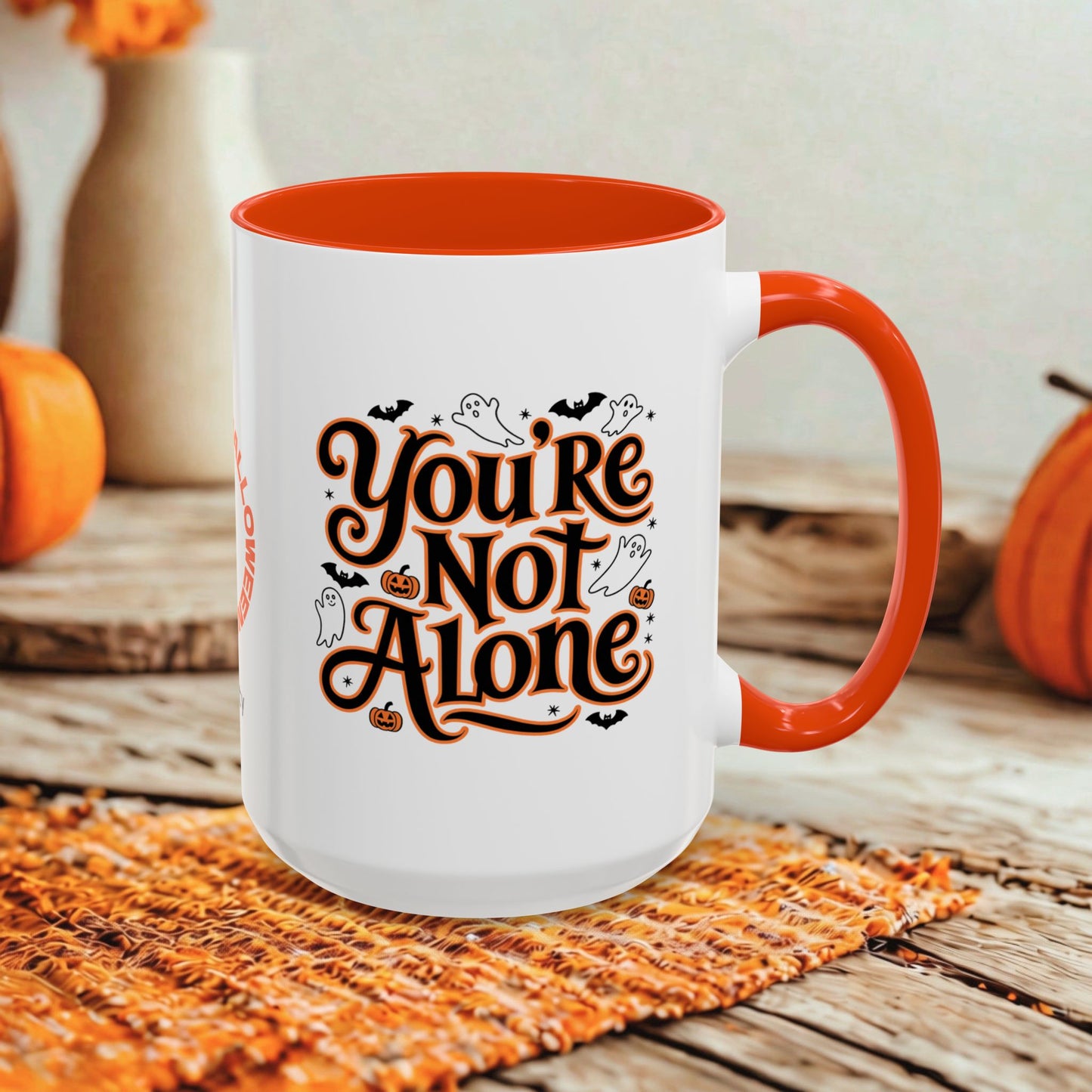 A white ceramic mug with a bright orange interior and handle, featuring the phrase 'You're Not Alone' in stylized brown lettering with black bats, ghosts, and pumpkins. The mug is set on a woven orange placemat on a rustic wooden table, with pumpkins and a blurred background.