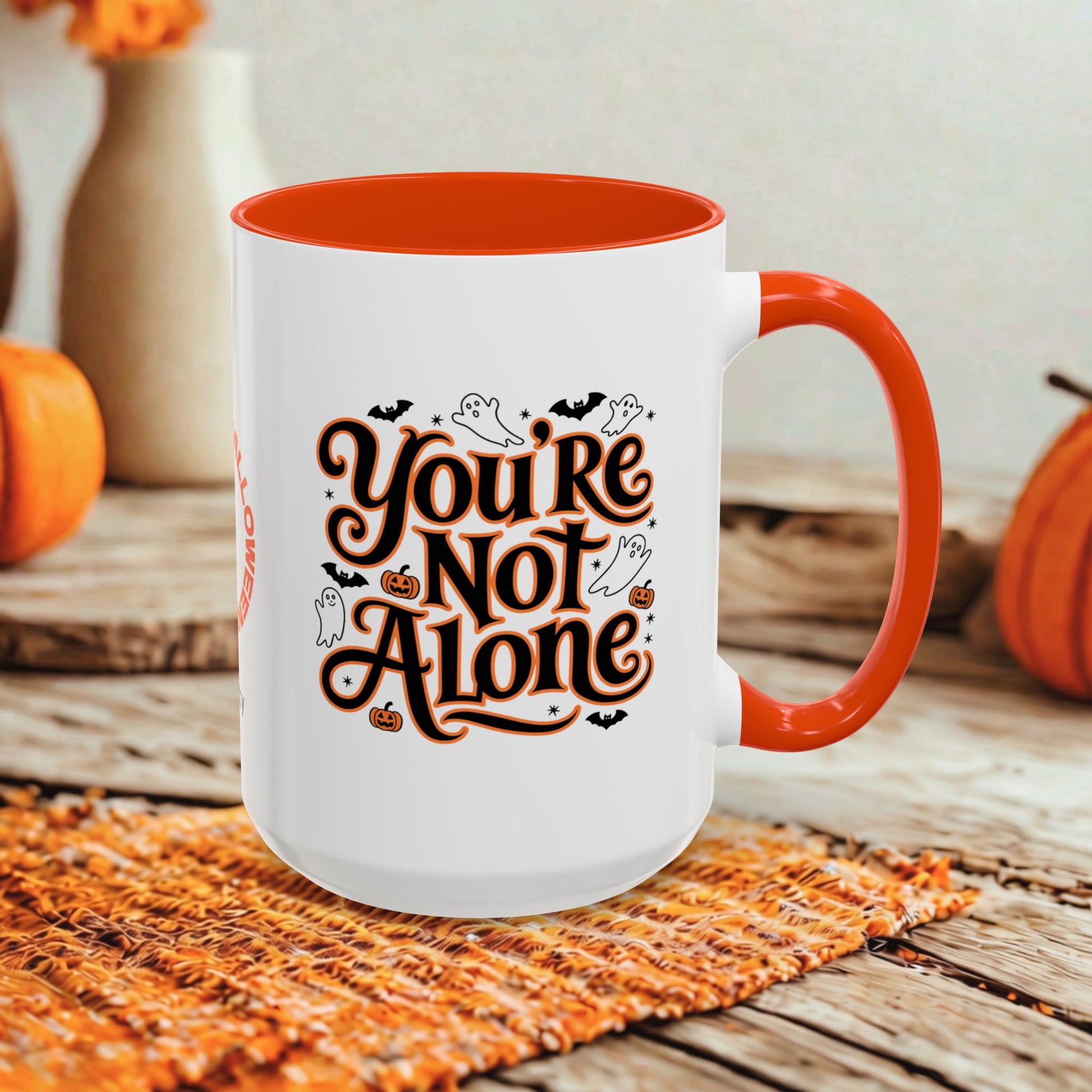 A white ceramic mug with a bright orange interior and handle, featuring the phrase 'You're Not Alone' in stylized brown lettering with black bats, ghosts, and pumpkins. The mug is set on a woven orange placemat on a rustic wooden table, with pumpkins and a blurred background.