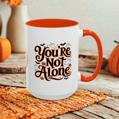 A white ceramic mug with a bright orange interior and handle, featuring the phrase 'You're Not Alone' in stylized brown lettering with black bats, ghosts, and pumpkins. The mug is set on a woven orange placemat on a rustic wooden table, with pumpkins and a blurred background.
