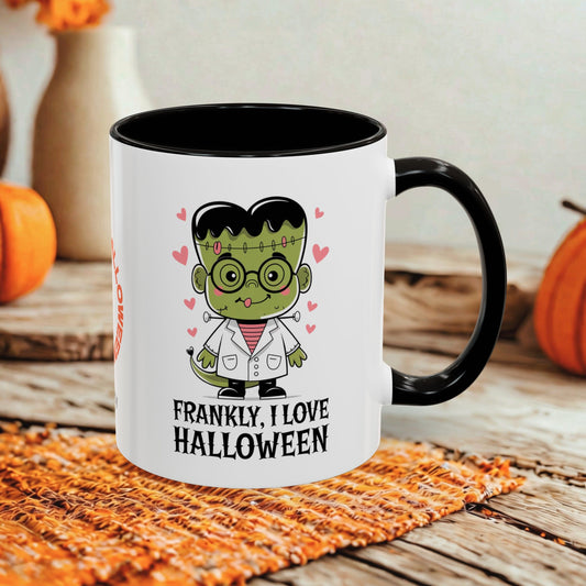 A white ceramic mug with a black handle and interior, featuring a cartoon Frankenstein character and the text 'FRANKLY, I LOVE HALLOWEEN' on the front. The mug is styled on a rustic wooden surface with a woven orange placemat and blurred pumpkins in the background.