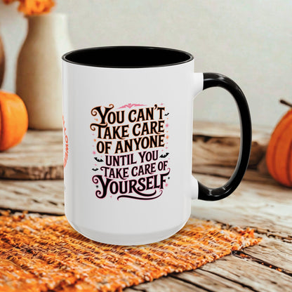 A white ceramic coffee mug with a black interior and black handle, featuring the printed phrase 'YOU CAN'T TAKE CARE OF ANYONE UNTIL YOU TAKE CARE OF YOURSELF' in dark brown and pink lettering. The mug is styled on a woven orange placemat on a rustic wooden table, with pumpkins and a blurred background.