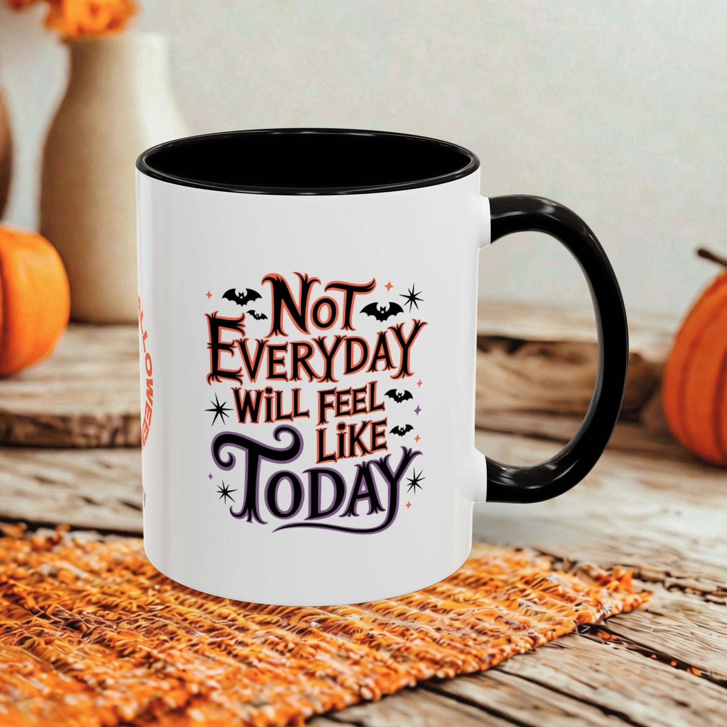 A white ceramic mug with a black handle and interior, printed with the phrase 'Not Everyday Will Feel Like Today' in stylized orange and purple lettering with small bat and star graphics. The mug sits on a woven orange placemat on a rustic wooden table, with pumpkins and a blurred background.