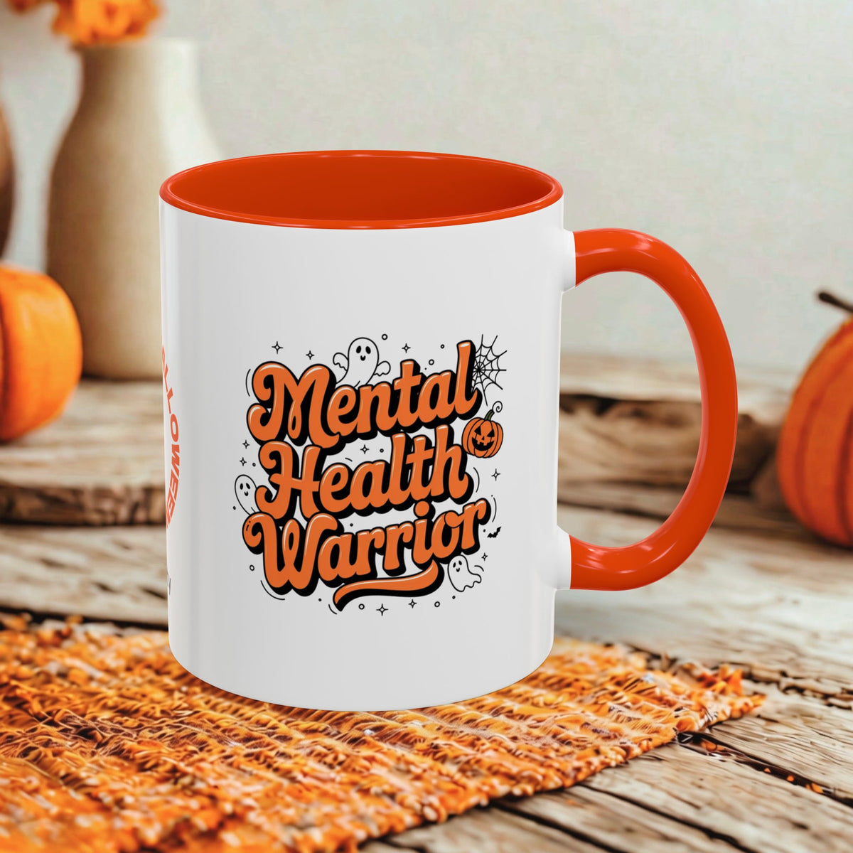 A white ceramic coffee mug with a bright orange handle and interior, featuring the text 'Mental Health Warrior' in orange and black with small Halloween-themed doodles. The mug is set on a woven orange placemat on a rustic wooden table, with decorative pumpkins and a neutral background.