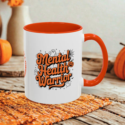 A white ceramic coffee mug with a bright orange handle and interior, featuring the text 'Mental Health Warrior' in orange and black with small Halloween-themed doodles. The mug is set on a woven orange placemat on a rustic wooden table, with decorative pumpkins and a neutral background.