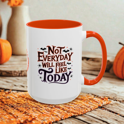 A white ceramic mug with a bright orange handle and interior, featuring the printed phrase 'Not Everyday Will Feel Like Today' in dark lettering with small bat and spider graphics. The mug is styled on a woven orange placemat over a rustic wooden surface, with pumpkins and blurred decorative vases in the background.