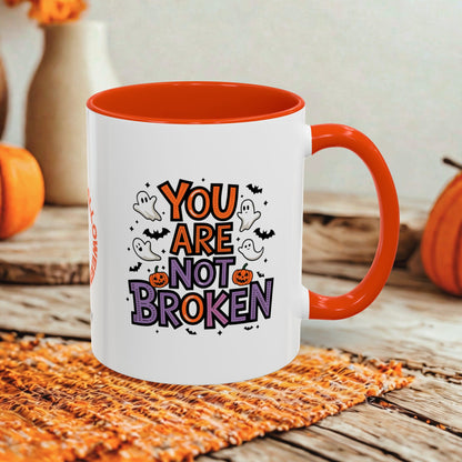 A white ceramic mug with a bright orange handle and interior, printed with the phrase 'YOU ARE NOT BROKEN' in orange, purple, and black lettering with cartoon ghosts, bats, and pumpkins. The mug sits on an orange woven placemat on a rustic wooden table, with blurred orange pumpkins and a neutral background.