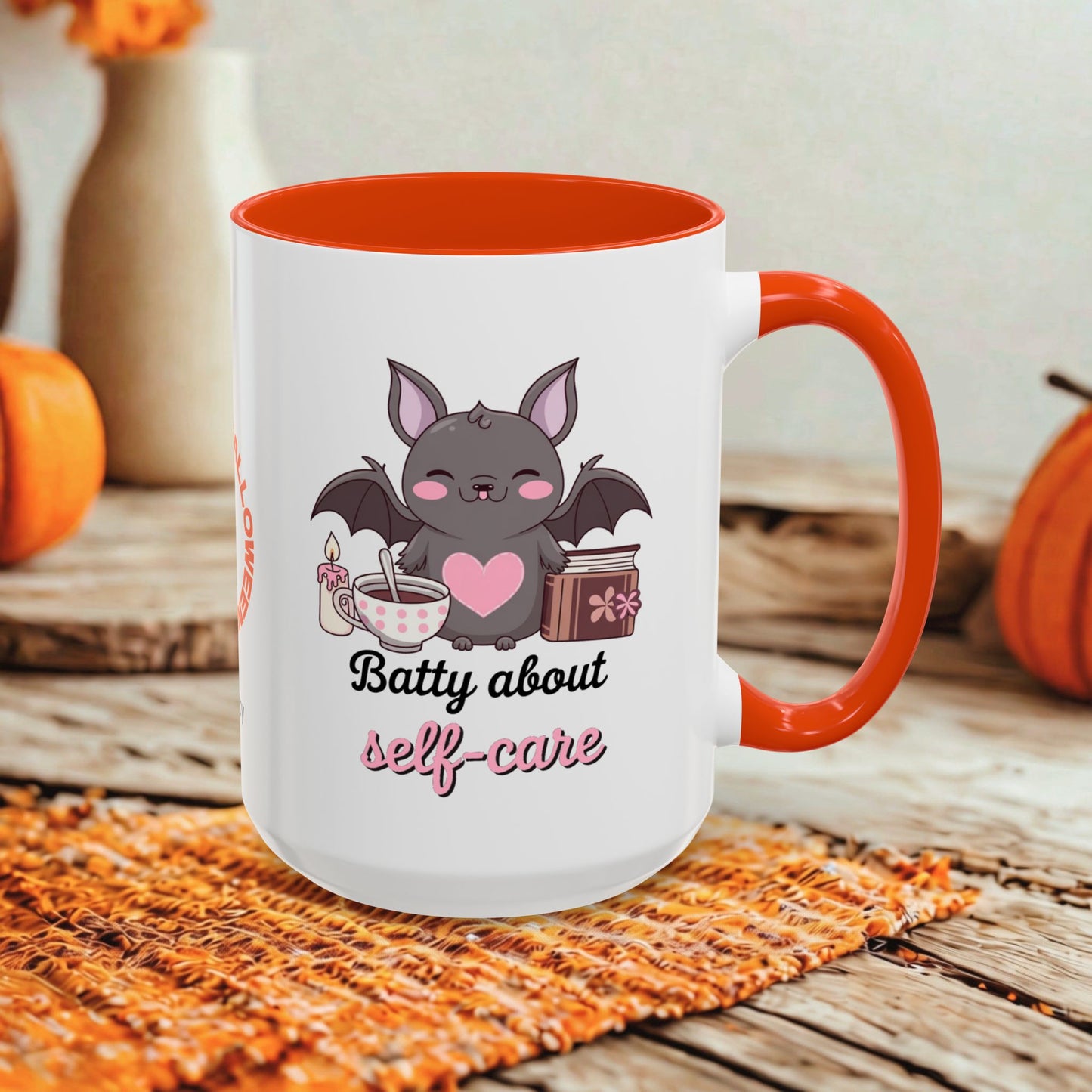 A white ceramic mug with a bright orange handle and interior, featuring a cute cartoon bat graphic and the text 'Batty about self-care' on the front. The mug is styled on a rustic wooden surface with autumn-themed props like pumpkins and a woven placemat.