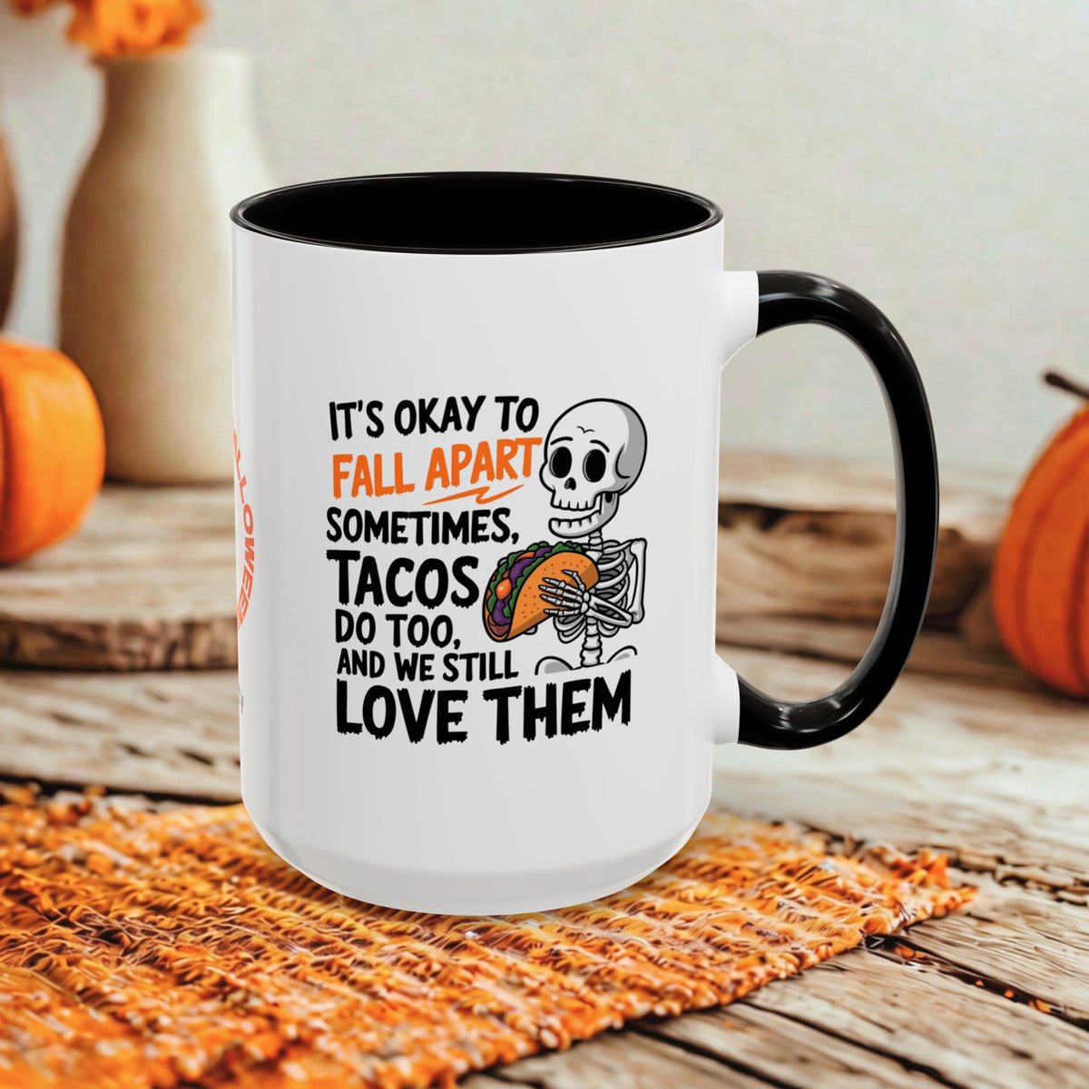 A white ceramic coffee mug with a black handle and black interior, featuring a humorous Halloween graphic with a cartoon skeleton and the text 'IT'S OKAY TO FALL APART SOMETIMES. TACOS DO TOO, AND WE STILL LOVE THEM.